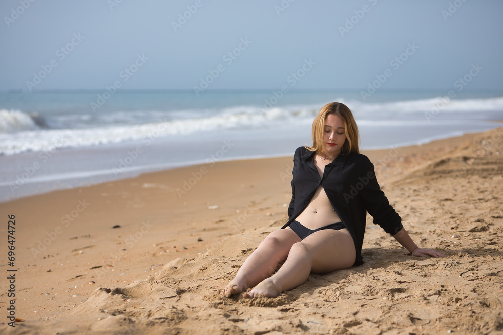 Young woman, blonde and beautiful, with a bikini and black shirt sitting on the beach, relaxed, quiet, peaceful, calm. Concept of solitude, relaxation, tranquility, peace.