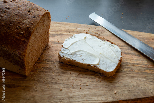 Slice of bread with mayonnaise spread on a wooden cutting board with bread next to it and a cutting knife