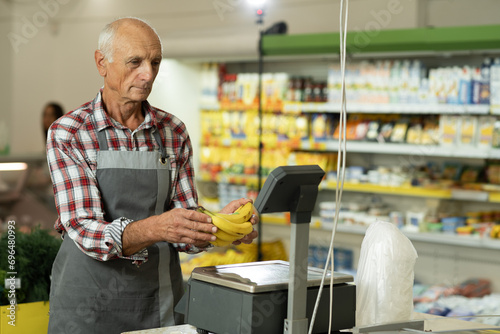 Old male salesman in a store weighing bananas on scales packing fruits waiting for a sale