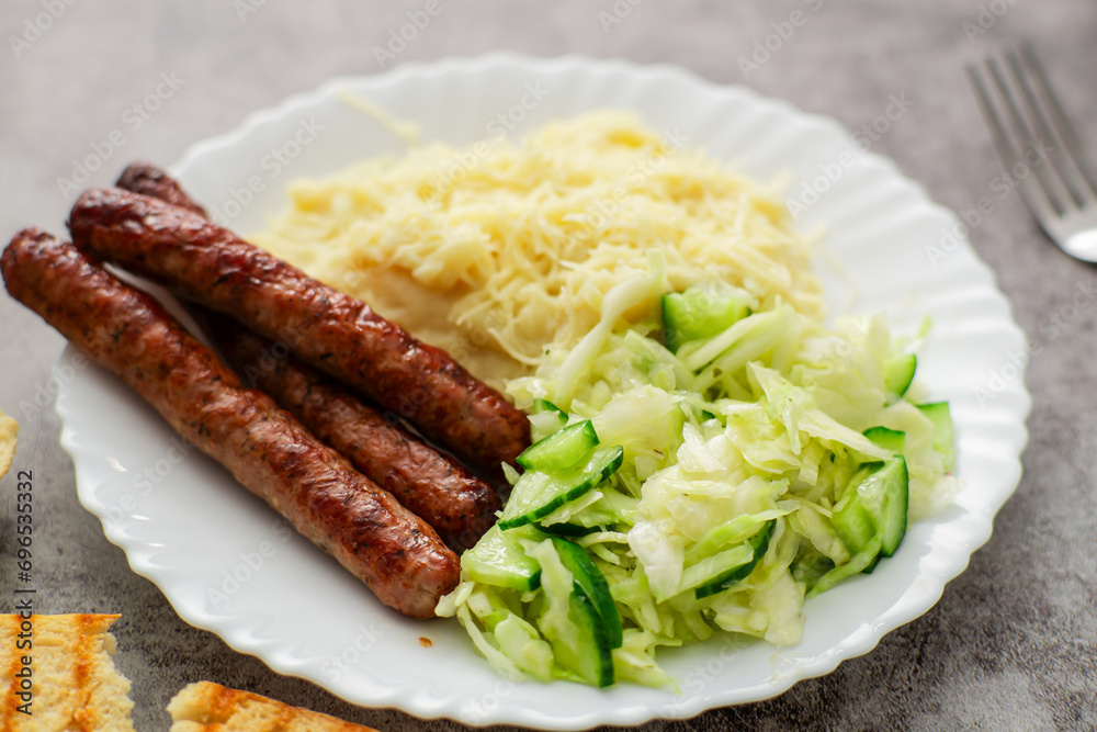 Enjoy the deliciousness of cheesy mashed potatoes, a fresh cabbage-cucumber salad, and mouthwatering sausages, all presented on a white plate against a dark surface