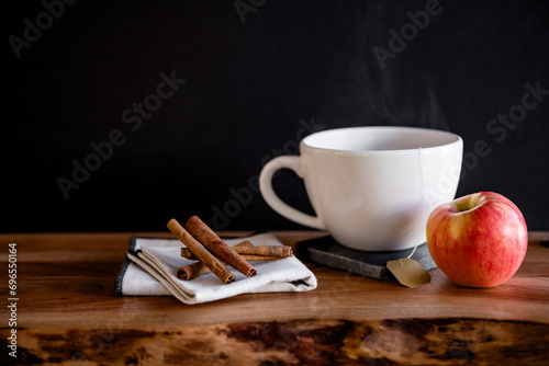 White tea mug with warm tea with apple slice and cinnamon sticks on wooden table with mulling spices