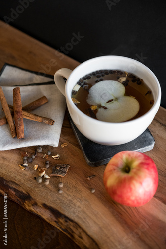 White tea mug with warm tea with apple slice and cinnamon sticks on wooden table with mulling spices