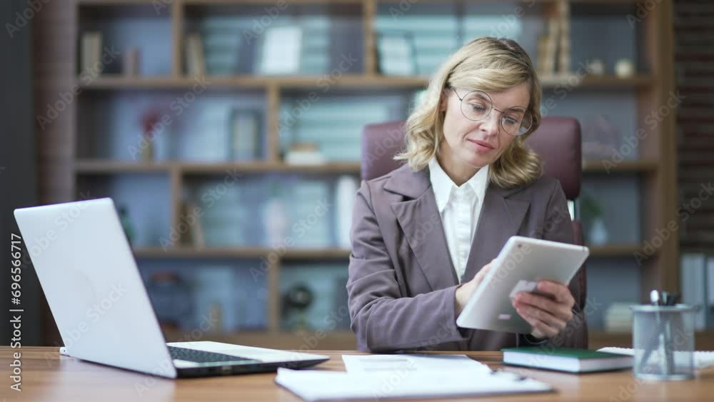 A businesswoman makes an accounting report using a laptop computer and digital tablet. Mature female accountant financier entrepreneur or small business owner doing paperwork with documents in office A businesswoman makes an accounting report using a laptop computer and digital tablet. Mature female accountant financier entrepreneur or small business owner doing paperwork with documents in office