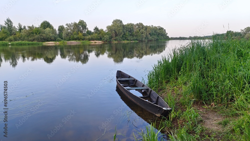 A wooden boat with benches moored to the grassy shore has gained water. There is aquatic vegetation in the water. The trees growing on the opposite sandy river bank are reflected in the water