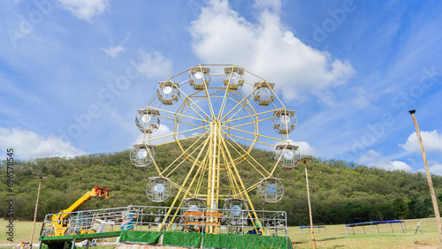 Location photos The large Ferris wheel is empty during the day. The backdrop is a calm, natural atmosphere. Green fields and big mountains, a clear sky, and yellow cranes and lampposts all around.