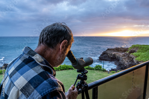 Mature Caucasian man by the ocean using a spotting scope to watch whales and dolphins as the sun setting over the ocean, Makahuena Point, Kauai, Hawaii photo