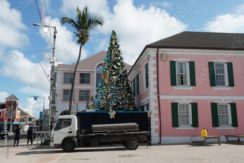 Christmas tree decorated with colorful balls in contrast with a palm tree on the Bahamas in Nassau port during the Christmas holidays. There are pink buildings and lorry.