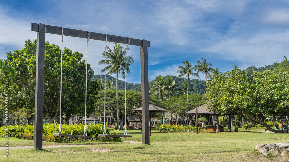 A recreation area in a tropical park. A wooden swing is suspended from a crossbar on ropes. Gazebos and canopies are visible among the green vegetation. Clouds in the blue sky. Malaysia. Borneo. 