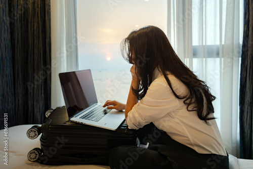 Asian woman packing suitcase on bed prepare for her travel and planning vacation travel with laptop, reading tourist blog online, booking tickets or hotel room on web getting ready for abroad journey.