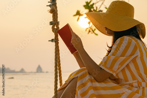 Outdoors lifestyle fashion portrait stunning asian woman enjoying on swing on the tropical island and reading book. In the background the sea and beautiful sunset.