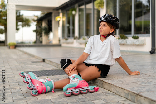 Positive teen girl in casual clothes smiling and looking at camera while sitting on asphalt road in roller skates