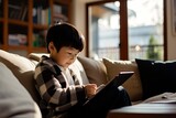 A young Asian boy using a tablet for education and entertainment, sitting on a sofa at home.