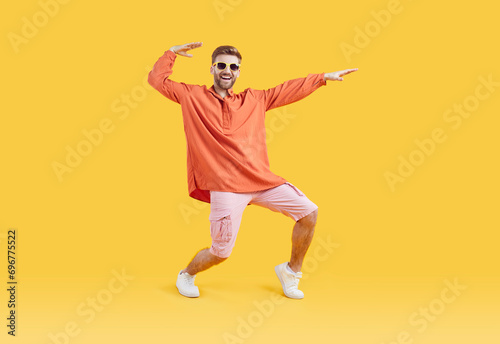 Happy guy in casual summer clothes having fun in fashion studio. Full body length shot of funny young man in orange shirt, pink shorts, white sneakers and sunglasses dancing on yellow color background photo