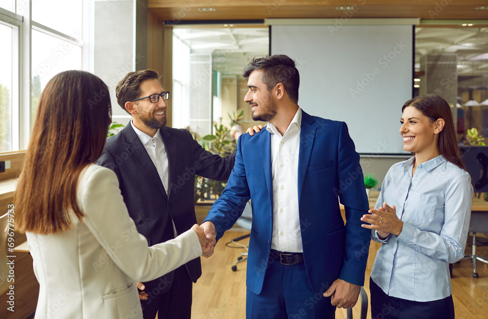 Woman shaking hands with a man celebrating success, making deal, business achievement, signing contract or greeting new employee with group of people in background standing in office. - obrazy, fototapety, plakaty 