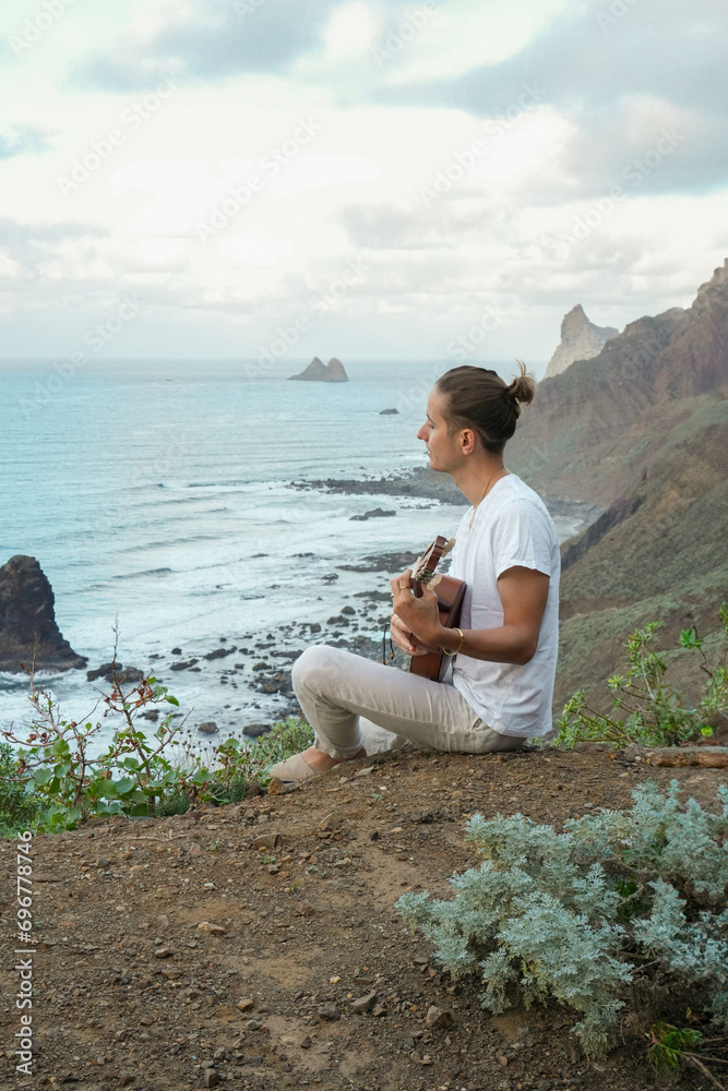 Young long hair man in casual attire, plays a ukulele on a cliff edge, overlooking a tranquil ocean and rugged cliffs