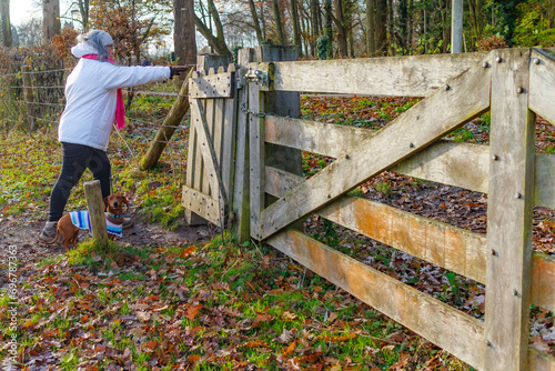 Adult female hiker leaving Dutch nature reserve Geleenbeekdal, opening gate, wooden fence, bare trees in misty background, white jacket, pink shawl, day in Schinnen, South Limburg, Netherlands photo