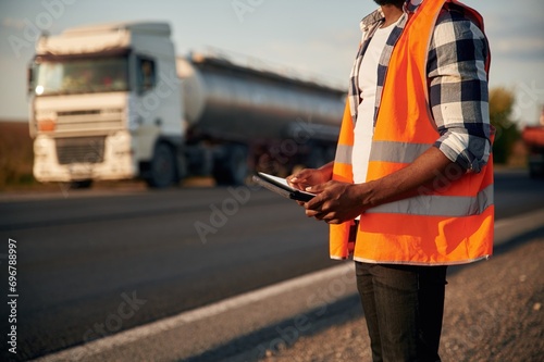 Close up view. Holding tablet. Black man is standing on the road with truck on it