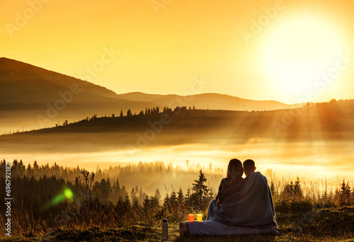a couple of a girl and a guy are sitting hugging and curling up with a blanket on a cliff overlooking the silhouettes of mountains in the fog in the dawn light. View from the back.