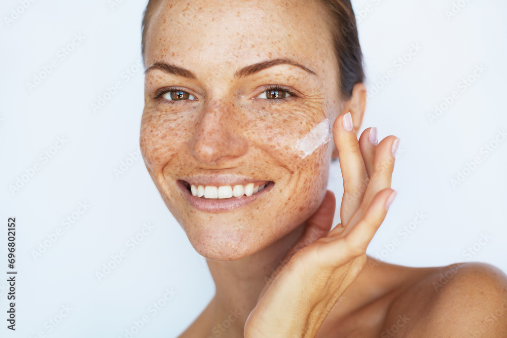Beautiful woman with a freckles is applying a facial skincare cream and smiling. Beauty skin care,  hydration and moisturising treatment - obrazy, fototapety, plakaty 