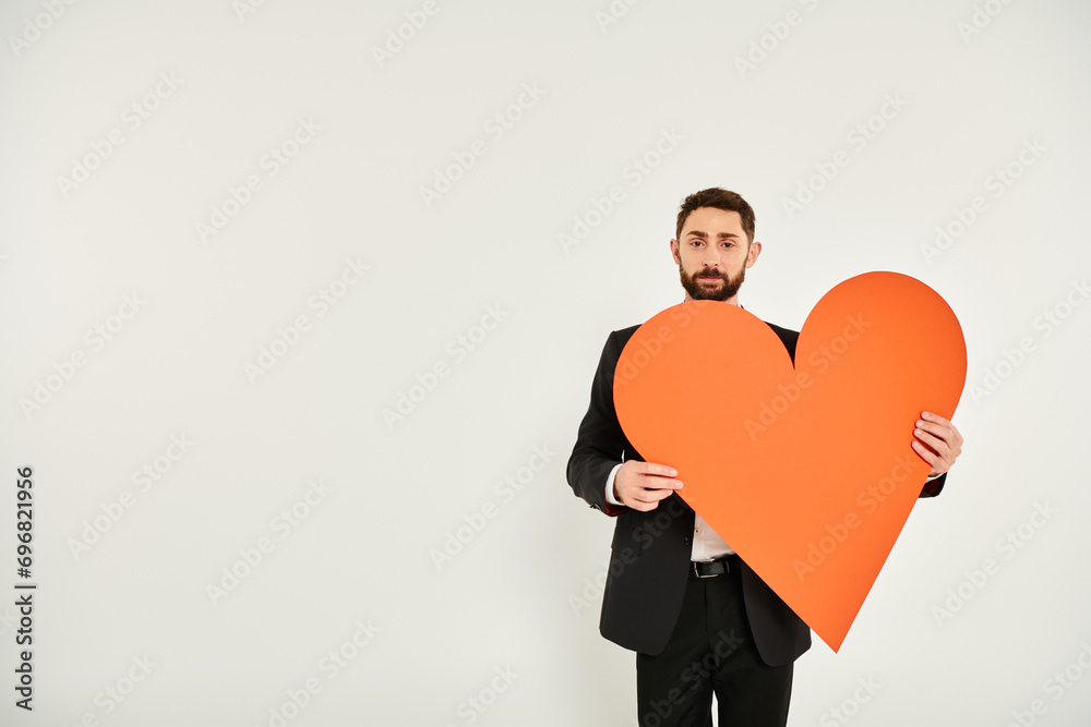 well-dressed bearded man with huge orange paper heart looking at camera on grey, st valentines day well-dressed bearded man with huge orange paper heart looking at camera on grey, st valentines day