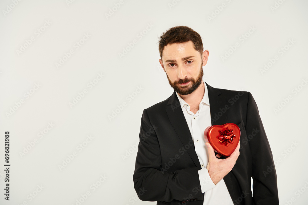 handsome elegant man with red heart-shaped Saint Valentines day present looking at camera on grey handsome elegant man with red heart-shaped Saint Valentines day present looking at camera on grey