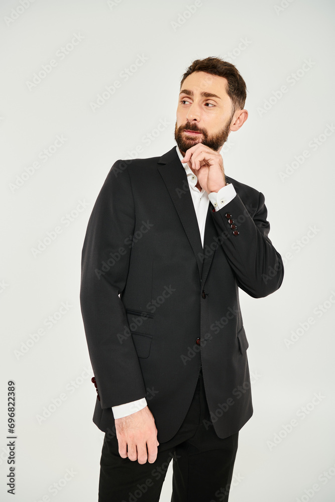 pensive elegant businessman in black suit touching beard and looking away on grey backdrop pensive elegant businessman in black suit touching beard and looking away on grey backdrop