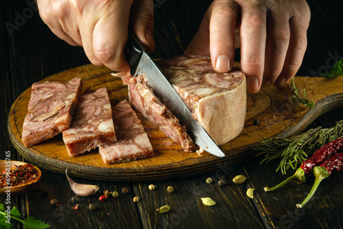 The cook cuts the brawn with a knife on the kitchen board before preparing sandwiches. Idea for a delicious snack with headcheese and aromatic spices. photo