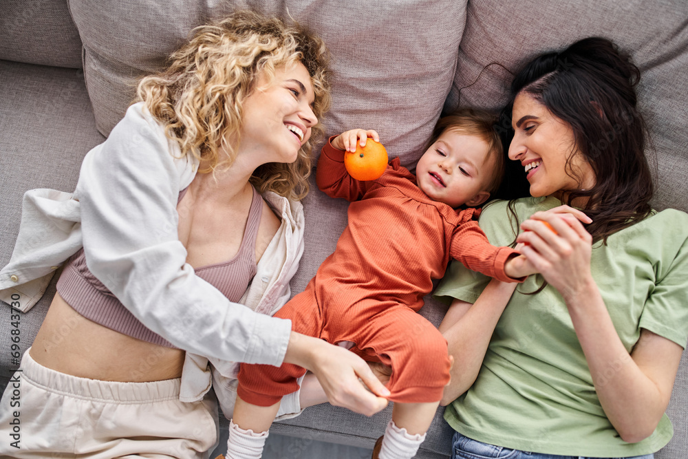 beautiful joyous lgbt couple lying on sofa with their baby girl holding tangerines, family concept beautiful joyous lgbt couple lying on sofa with their baby girl holding tangerines, family concept