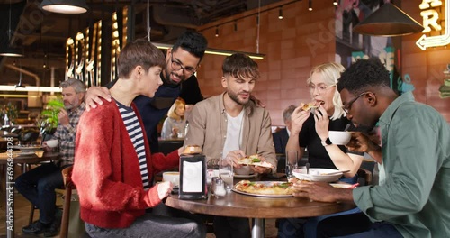 Company of four friends sitting at table and eating tasty Italian pizza in cafe. Young multiethnic waiter coming to his clients and communicating with them. People having meet in restaurant.