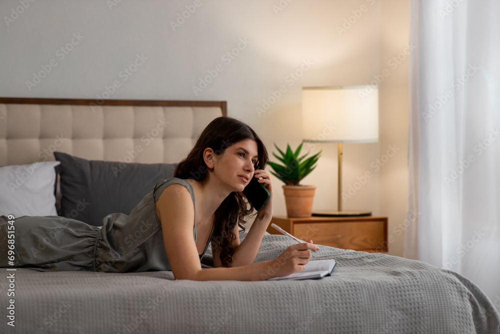 Pretty girl lying on the bed with diary and talking on the phone. Young woman with notebook and pen Pretty girl lying on the bed with diary and talking on the phone. Young woman with notebook and pen