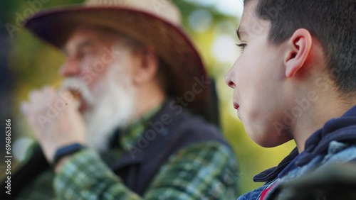 Concept of family vacation. Lunch together tradition. Grey bearded grandfather sitting in chair and telling life stories to his little grandson. Fishing with grandchild. Eating sandwiches in forest photo