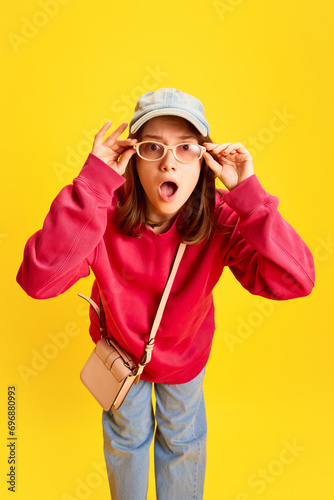Funny girl in fashion outfit holding sunglasses and confusing, surprised looking at camera against yellow studio background. Concept of fashion and style, traveling, culture, fun and joy.