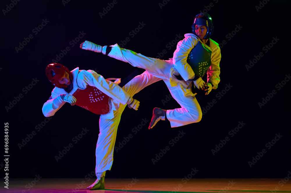 Dynamic image of young men, taekwondo athletes in kimono and helmets training against black background in neon light. Concept of martial arts, combat sport, competition, action, strength