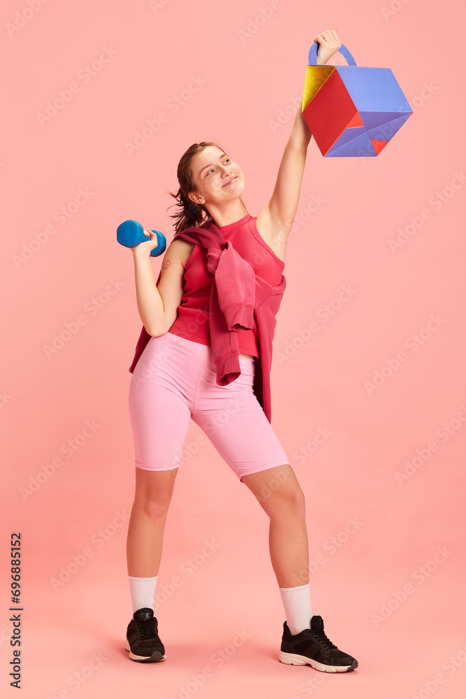 Full length portrait of athletic young lady training with dumbbells and paper pocket for delivery against pastel pink background. Concept of sport, active lifestyle, fitness, energy. Full length portrait of athletic young lady training with dumbbells and paper pocket for delivery against pastel pink background. Concept of sport, active lifestyle, fitness, energy.