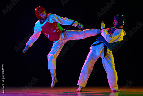 Dynamic image of young men, taekwondo athletes in kimono and helmets training against black background in neon light. Concept of martial arts, combat sport, competition, action, strength