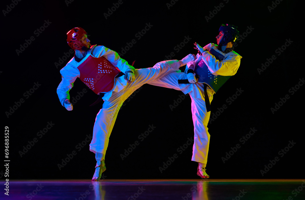 Competitive strong young men, taekwondo, karate athletes in motion, fighting, training against black background in neon light. Concept of martial arts, combat sport, competition, action, strength