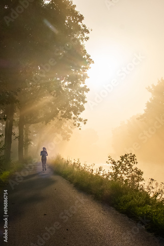 An early morning runner takes to a quiet country road, enveloped in a soft, golden mist. The sun, diffused through the fog, casts a warm, ethereal glow and creates striking rays of light through the