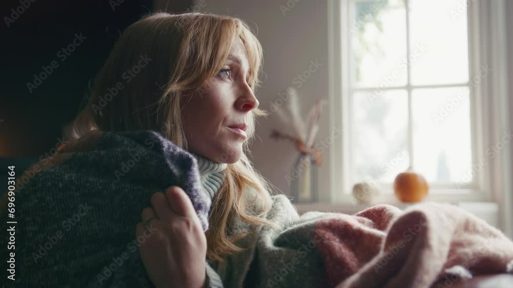 Head and shoulders shot of unhappy woman wearing cosy jumper and blanket sitting on sofa at home looking thoughtfully out of window - shot in slow motion