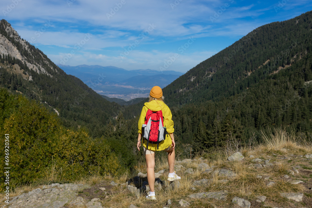 A girl on top of a mountain with green grass looks at a beautiful mountain valley. Landscape with a sporty young woman in a yellow raincoat. Travel and tourism. Hiking