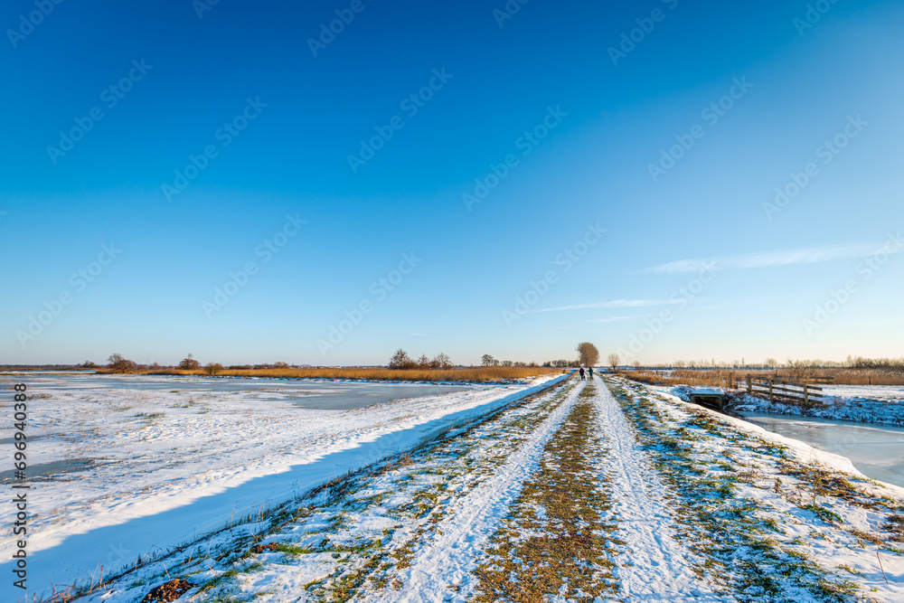 Dutch polder landscape on a sunny afternoon in the winter season. At the end of a long unpaved road two women are walking with their dog. Dutch polder landscape on a sunny afternoon in the winter season. At the end of a long unpaved road two women are walking with their dog.