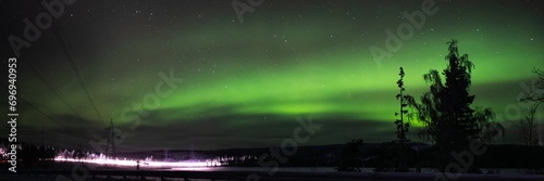 Panoramic night view of vibrant aurora borealis over a snowy landscape with trees and power lines, illustrating the natural phenomenon and arctic environment