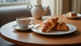  two croissants on a plate next to a cup of coffee and a teapot on a table with a teapot and a window in the background.