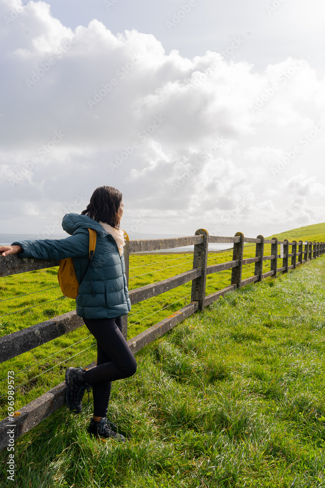 Woman with backpack enjoying the views leaning on a fence that disappers at the end of the green Irish landscape