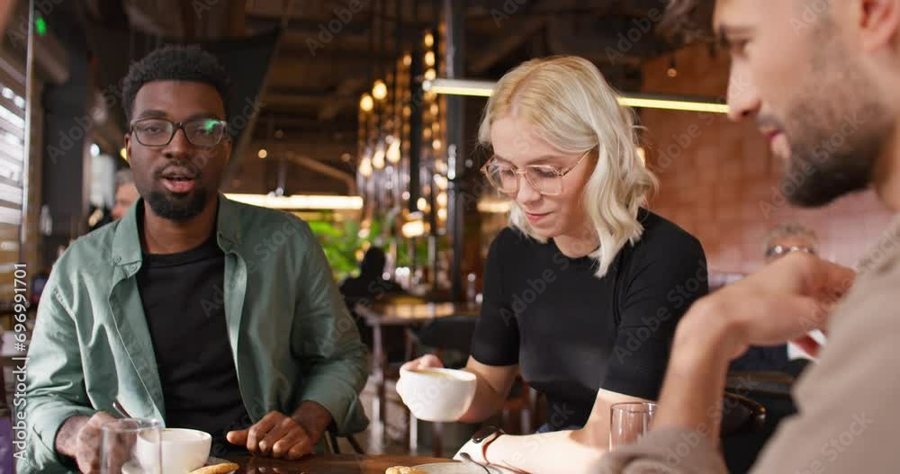Camera view of mixed-raced friends sitting together at table in cafe. Actively speaking with each other and happily smiling. Having positive conversation. People resting in bar at weekend.