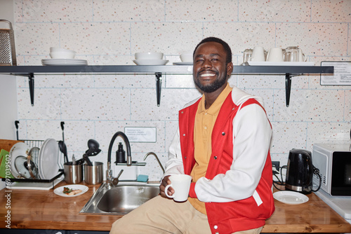 Waist up portrait of smiling Black man wearing varsity jacket smiling at camera in kitchen, with flash photo