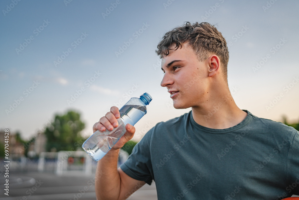 caucasian man teenager open plastic bottle of water outdoor in sunny day drink while hold basketball