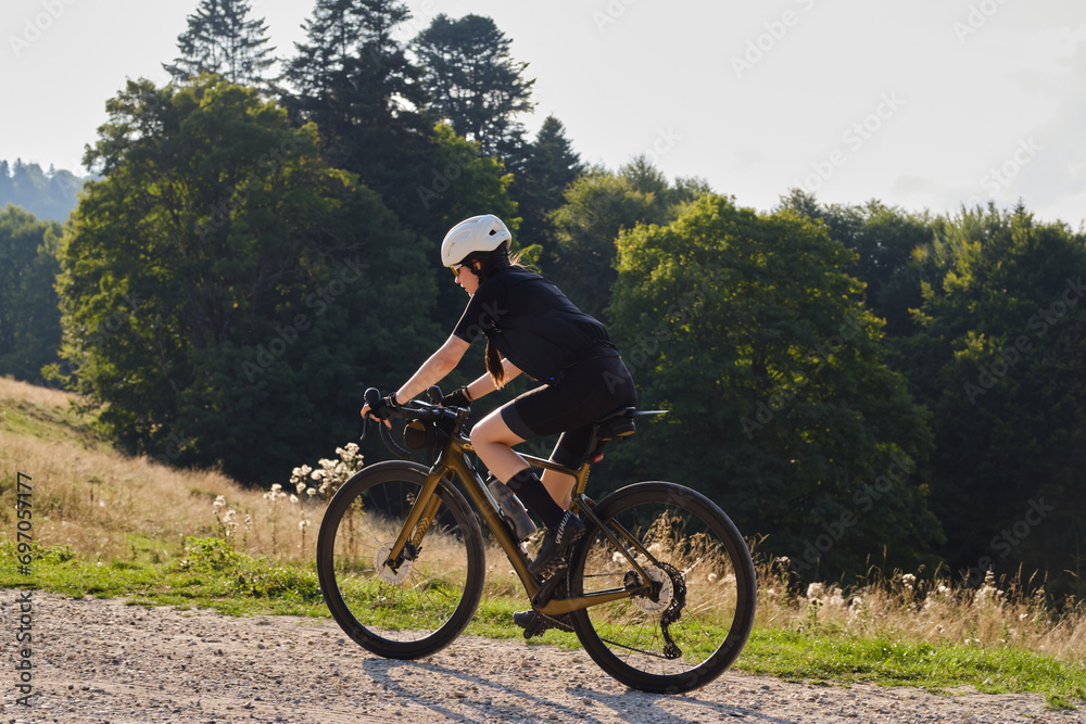 Woman cyclist, dressed in a black cycling kit and a white helmet,is riding a gravel bicycle on a gravel road with beautiful Romanian mountains in the background.Cycling adventure. Bucegi Natural Park.