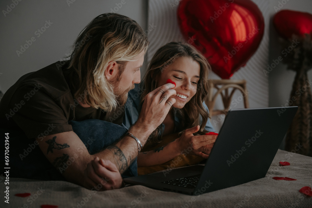 Happy loving couple shopping online while lying in bed and using laptop with heart shape balloons Happy loving couple shopping online while lying in bed and using laptop with heart shape balloons