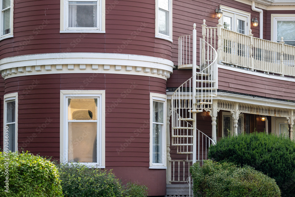 A street view of a spiral staircase on the outside of a large burgundy colored wooden house with multiple closed glass windows. Lush green shrubs are in front of the historic circular building. 