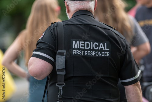 A close-up of a Caucasian male emergency health medical first responder or paramedic wearing a black uniform with grey letters. The ambulance attendant is wearing a short-sleeve shirt with lettering.